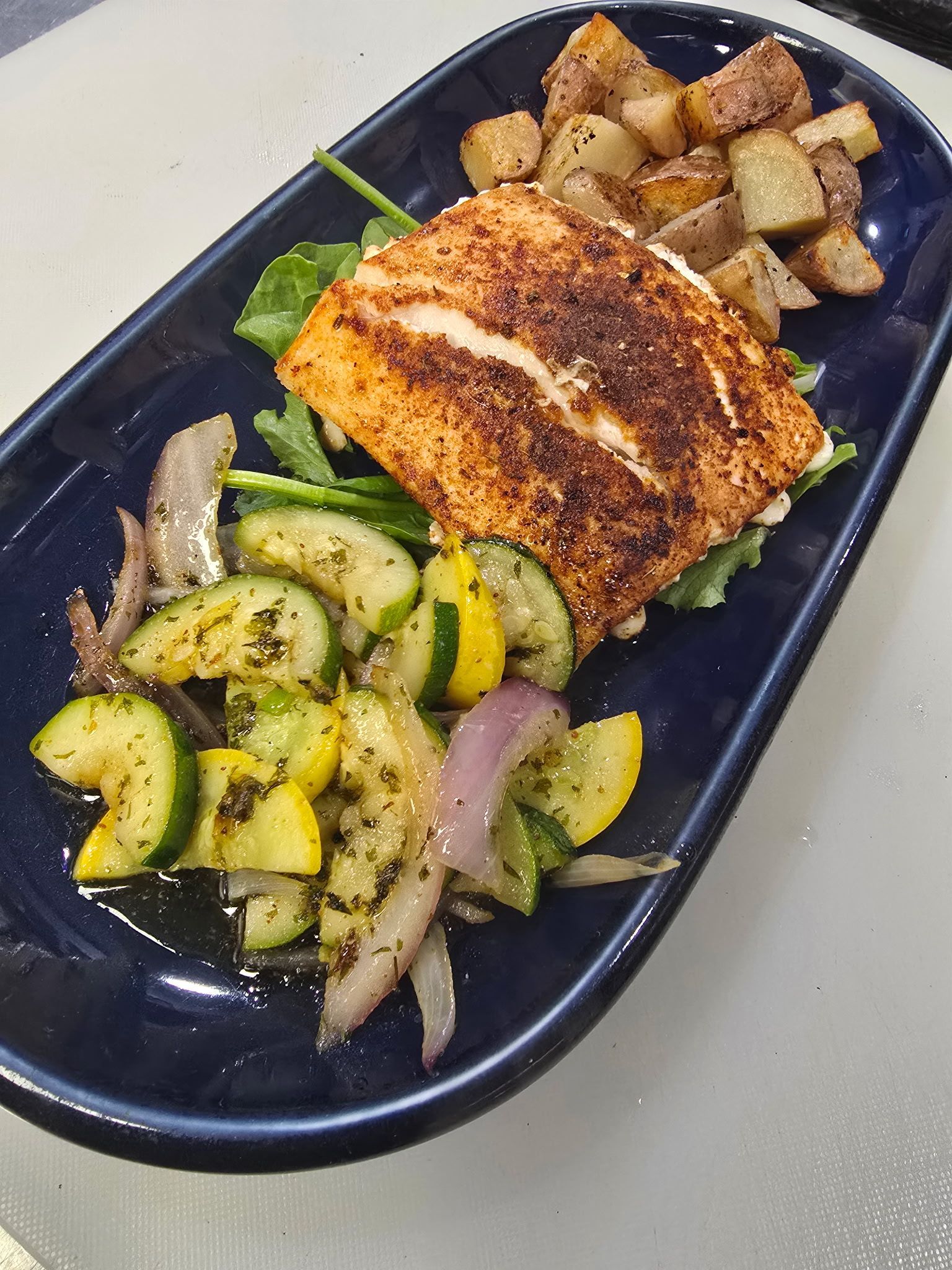 Plated grilled salmon with roasted potatoes and sautéed zucchini and onions on a dark oval dish
