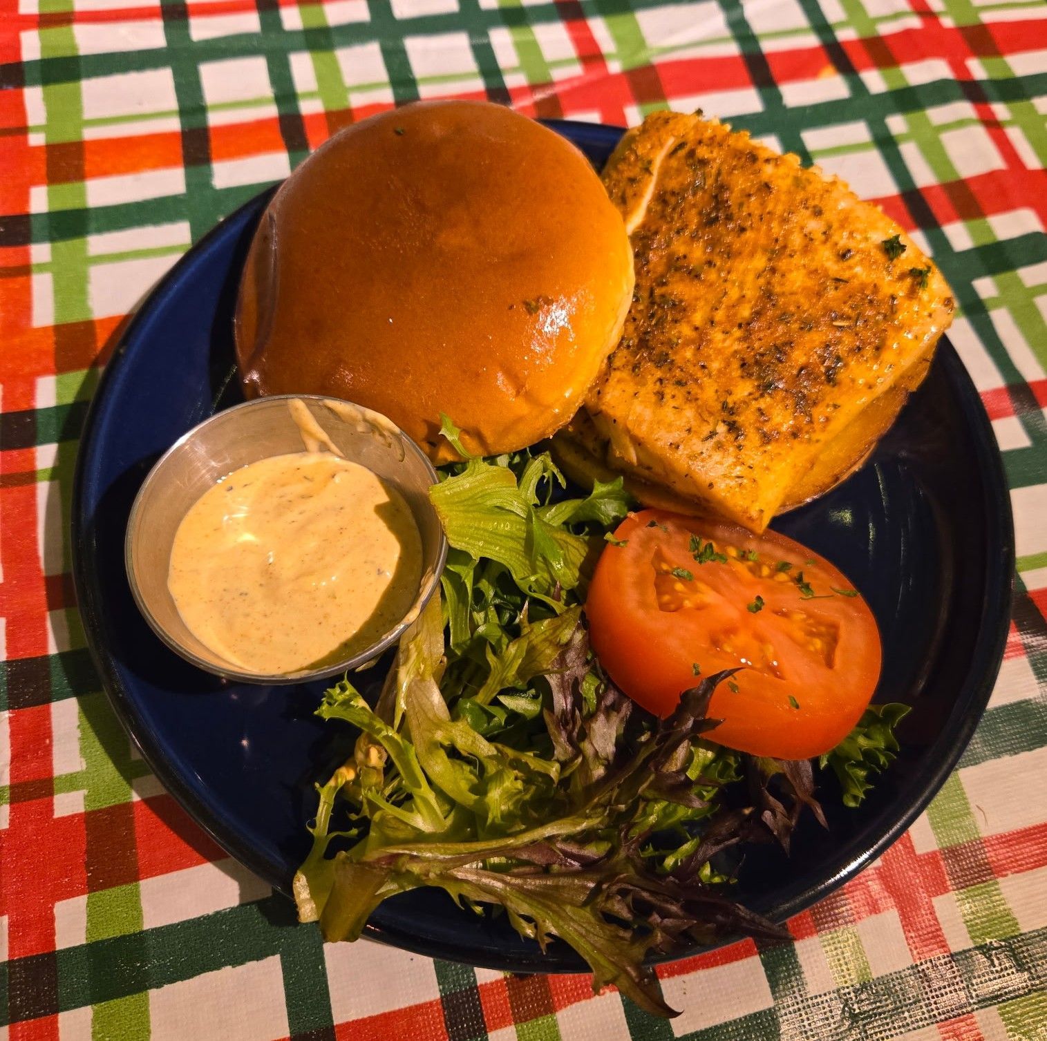 Mahi burger on a blue plate with sauce, tomato, and greens, on a checkered table.