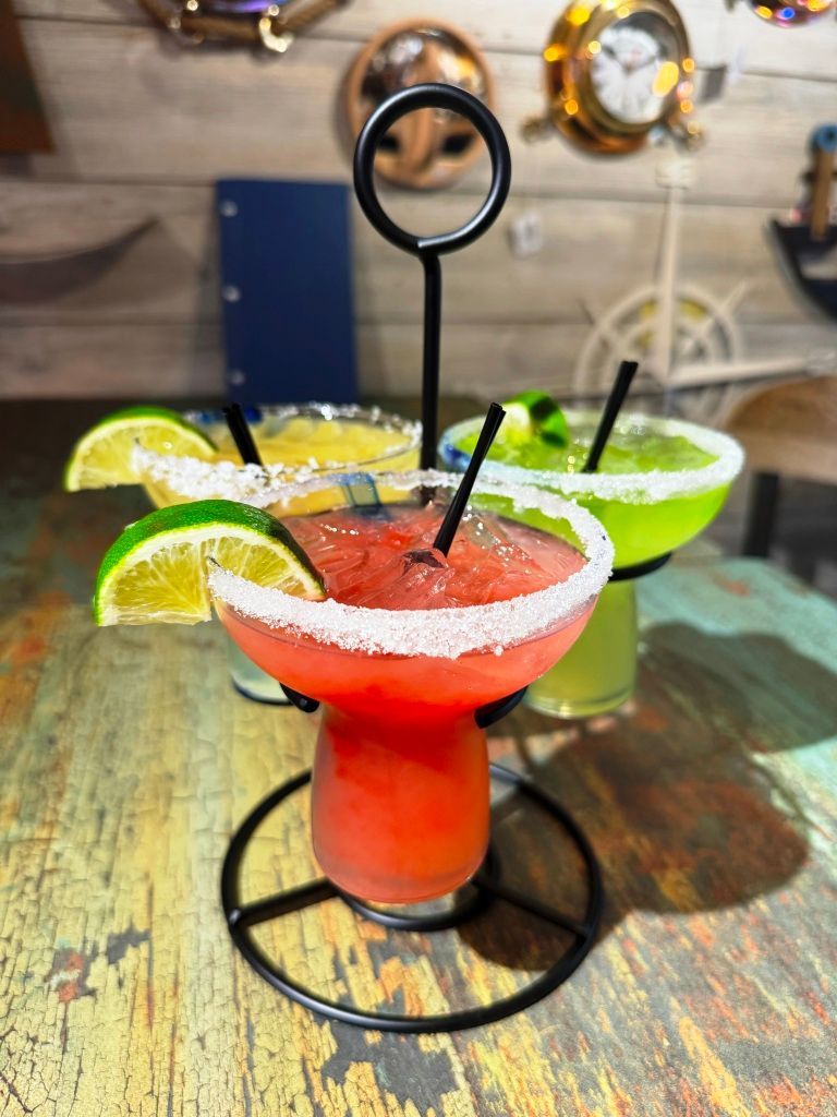 Three colorful margaritas with salt rims, in a tiered stand on a distressed wooden table, nautical themed decor.