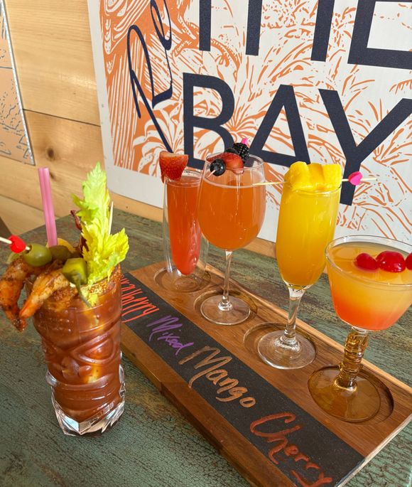 A flight of cocktails on a wooden tray, including a Bloody Mary and fruity drinks.