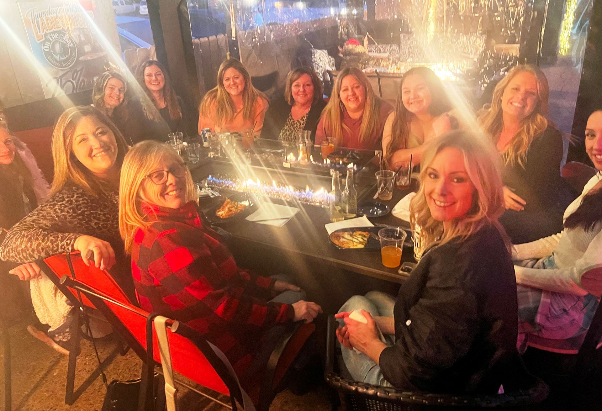 Group of women smiling around a fire pit at an outdoor restaurant, enjoying drinks and conversation.