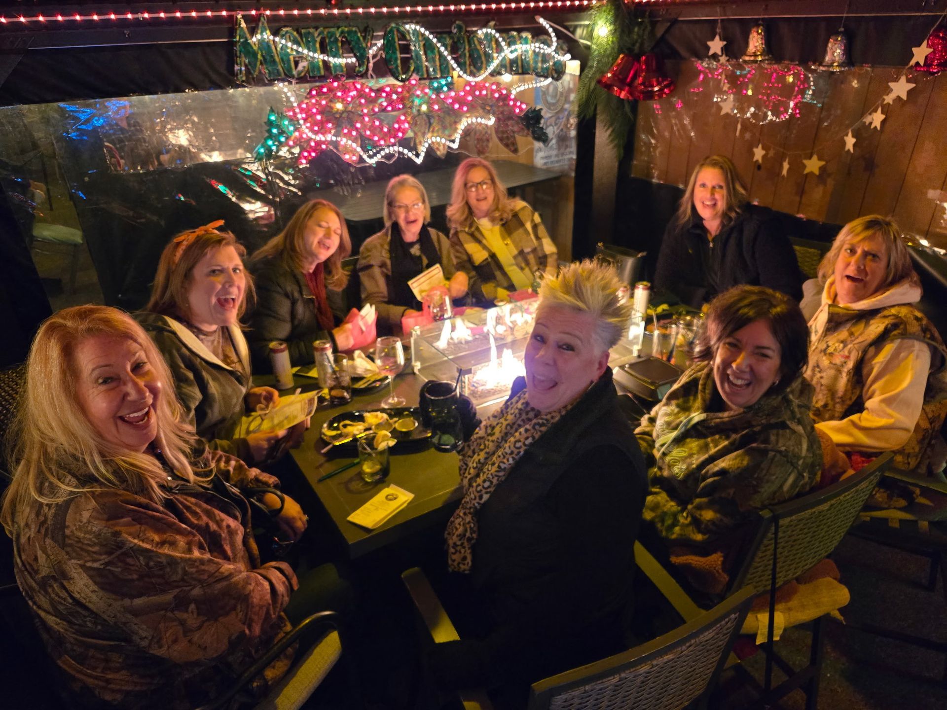 Group of women smiling, gathered around a lit outdoor table; festive lighting in the background.