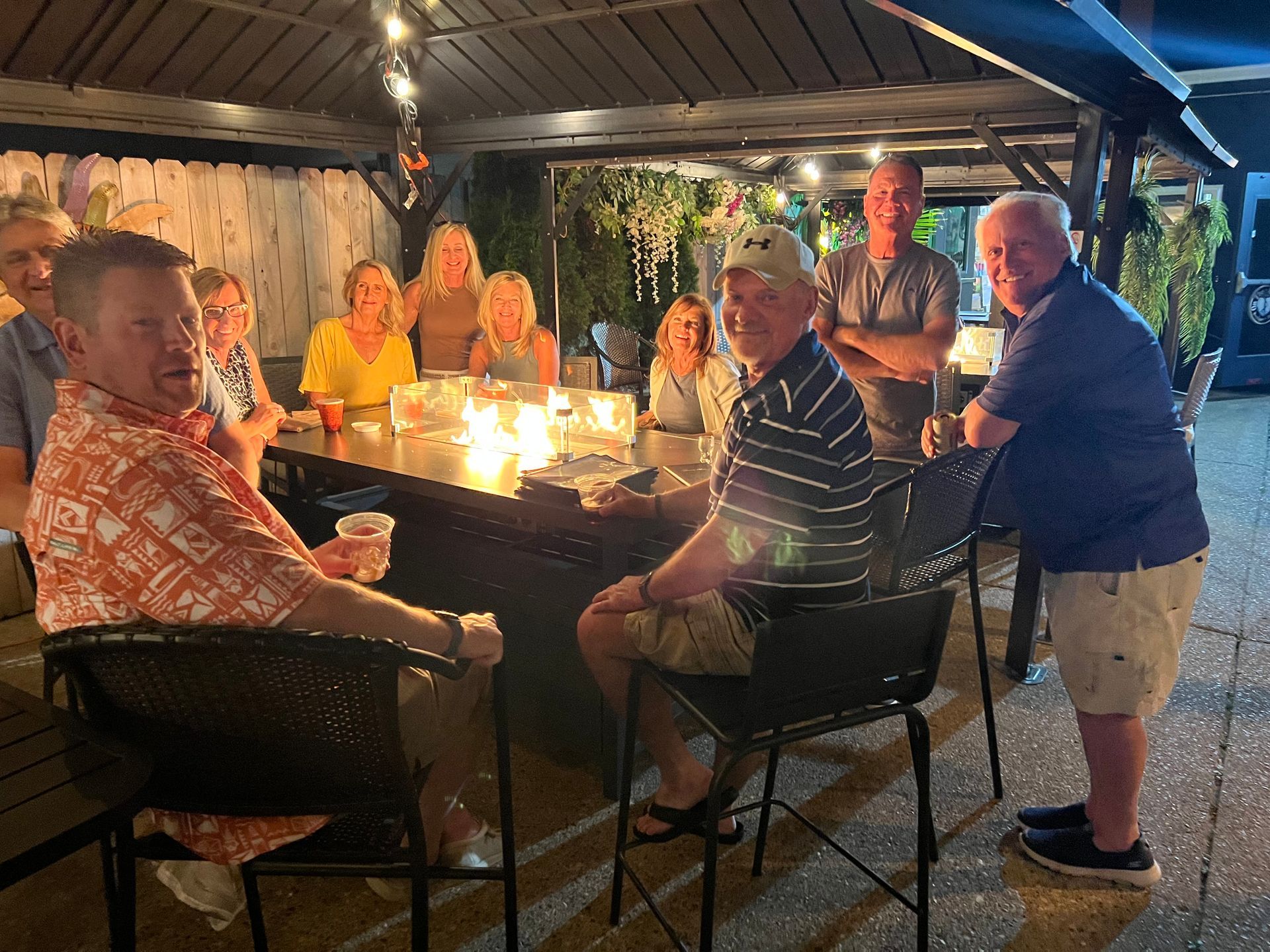 People gather around a fire pit in an outdoor patio. Some are seated, others standing, enjoying the evening.