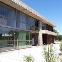 Edificio moderno con fachada de cristal, techo inclinado y pasarela ajardinada, bañado por la luz del día.