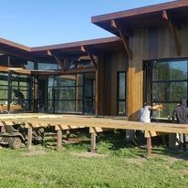 Casa moderna con grandes ventanales y una terraza de madera en un jardín con césped bajo un cielo azul.