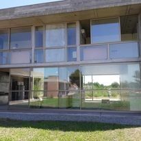 Edificio moderno con fachada de cristal, grandes ventanales, una pasarela cubierta y césped en la parte delantera.