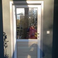 Puerta de entrada de cristal con marco blanco y reflejo de una persona vestida de rosa sosteniendo la cámara.