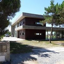 Edificio moderno de dos plantas con paredes de ladrillo oscuro, grandes ventanales y árboles a lo largo de la entrada.
