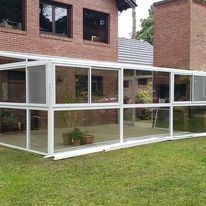 Terraza acristalada con marco blanco, adosada a una casa de ladrillo, situada sobre un césped.