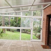 Terraza acristalada con paredes de cristal, suelo de baldosas y vistas a un jardín verde exterior.