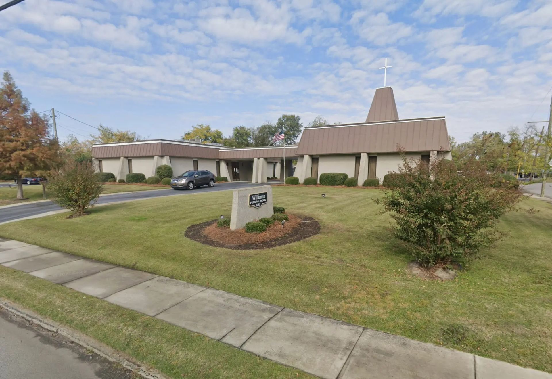 Suburban office building with beige walls, sloped roof, lawn, sidewalk, and parked cars under a cloudy sky