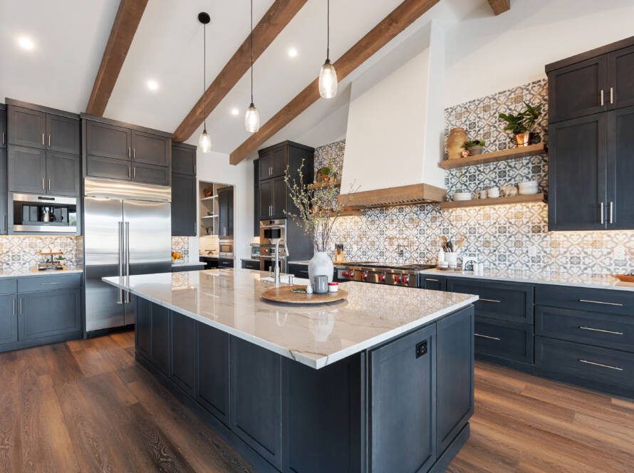 A modern kitchen with dark blue cabinets, a large white marble island, wood ceiling beams, and patterned tile backsplash.