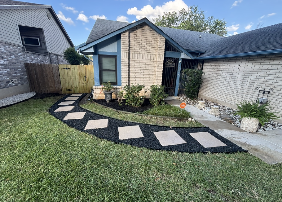 A front yard landscape featuring a curved stone paver walkway set in black mulch leading to a brick house entrance.