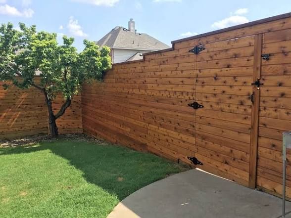 A stained horizontal wood fence with a gate, hinges, and a tree in a residential backyard with grass and a concrete patio.