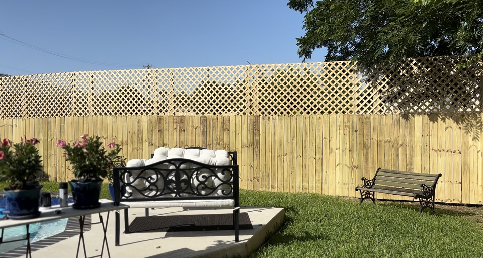 A wooden fence with a lattice top border stands behind a patio featuring a bench, potted plants, and a small metal bench.