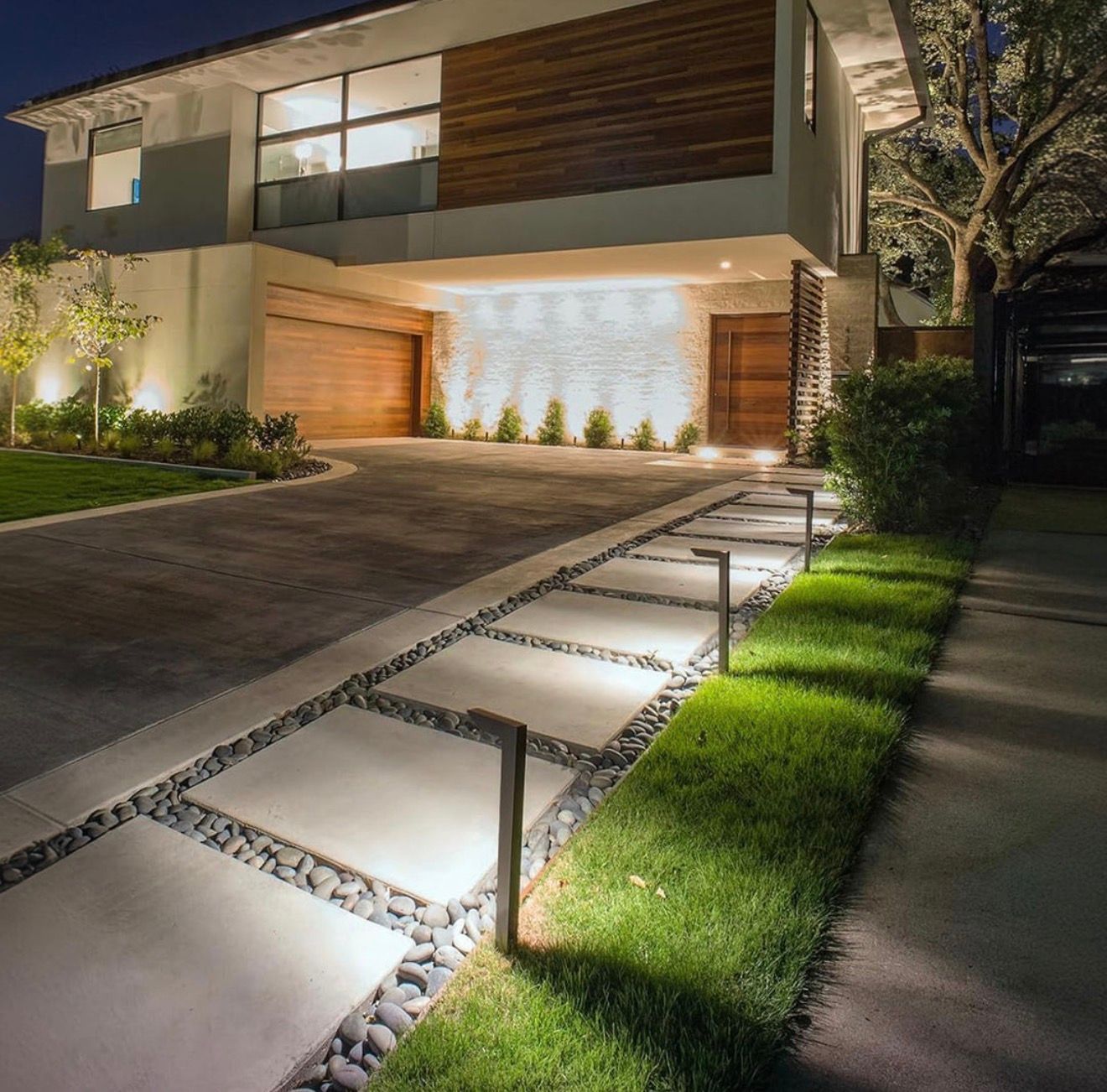 A modern house exterior at night, featuring a lit concrete paver pathway with gravel borders and warm wall lighting.