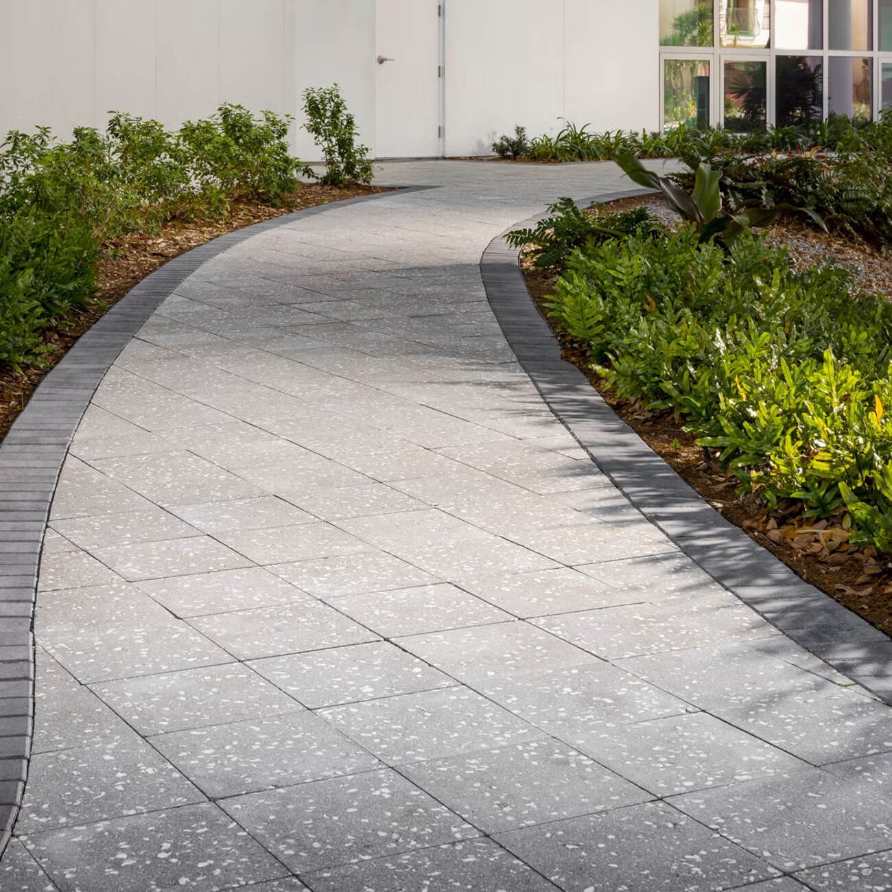 A winding gray paved path framed by dark stone borders, flanked by lush green bushes leading toward a white building.