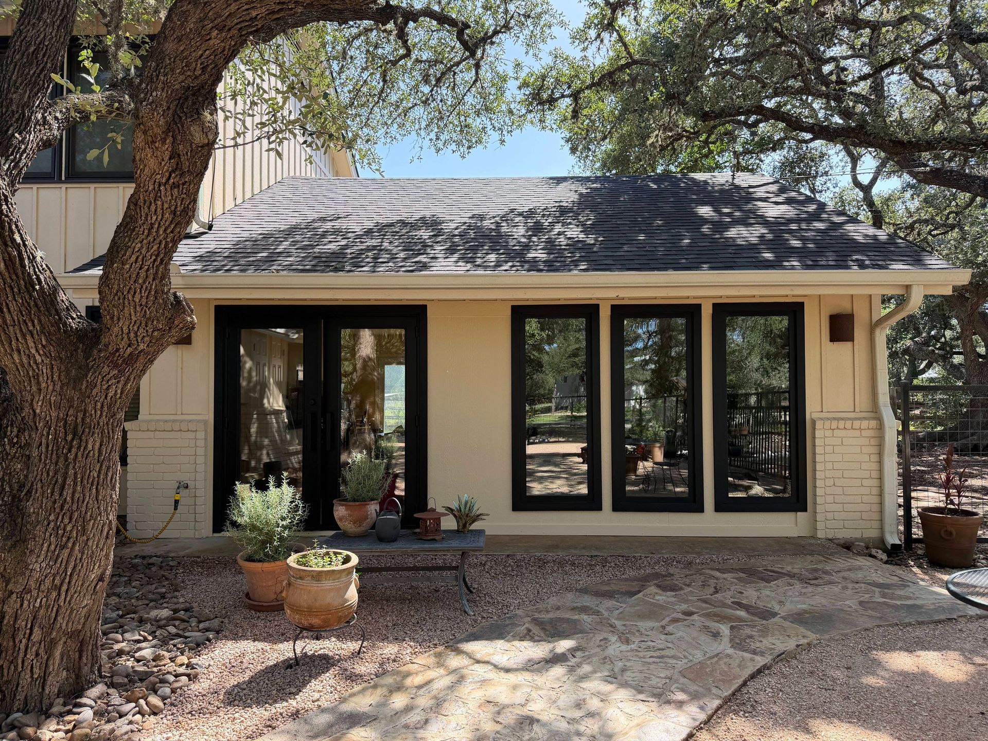 House exterior with black-framed glass doors, a shaded patio, and large trees in front