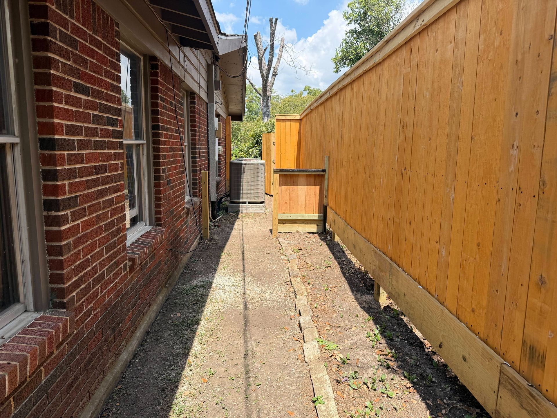 Narrow gravel alley between a red brick house and a tall wooden fence, with a small gate ahead.