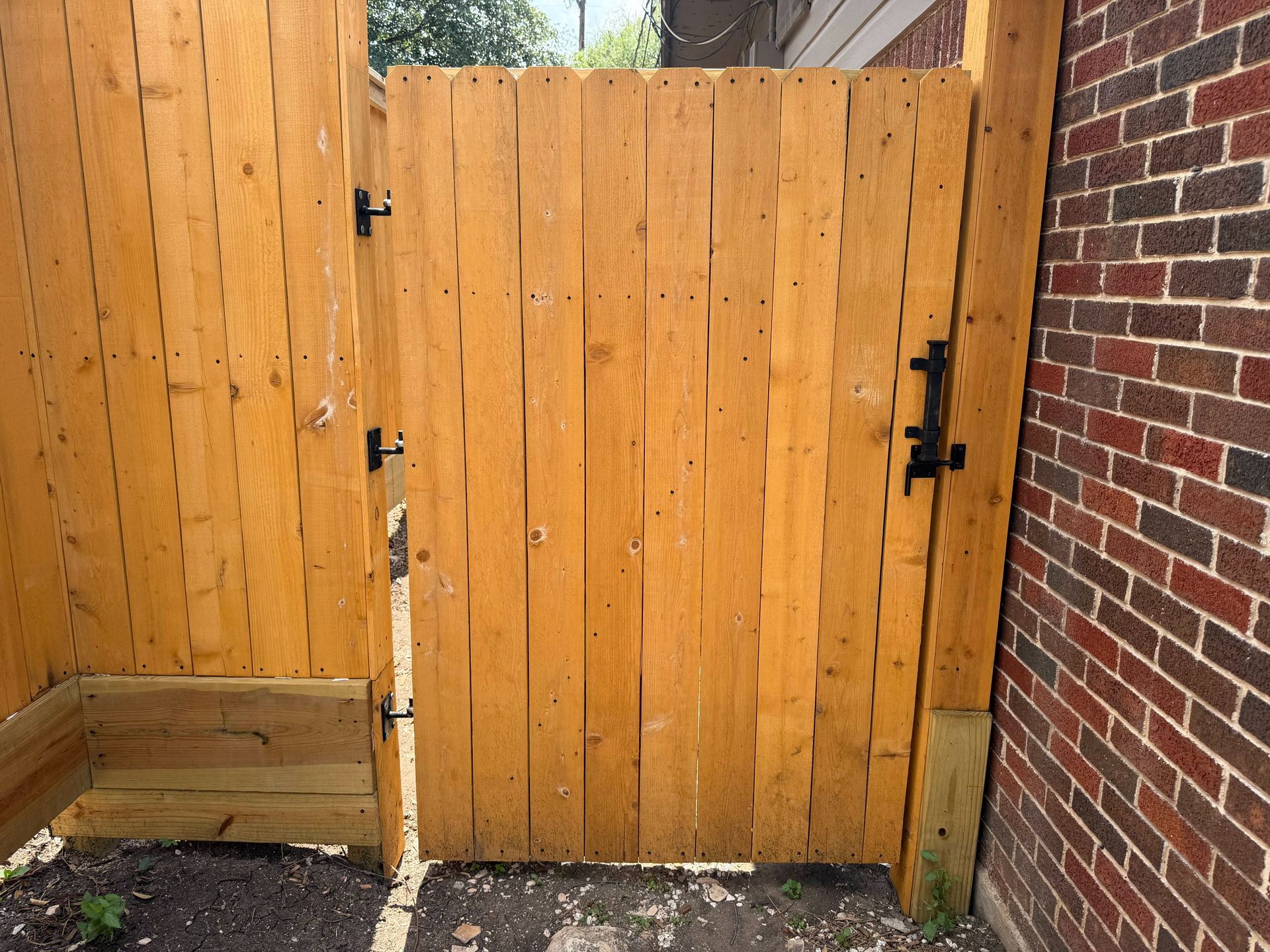 Wooden backyard gate beside a brick wall and fence, with black latches and gravel below.