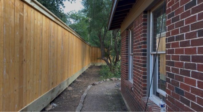 Narrow side yard between a wooden fence and a brick house, with a dirt path and trees ahead.