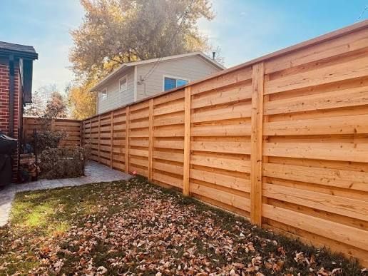 A long, horizontal-slat natural wood fence in a backyard, partially covered in fallen autumn leaves.