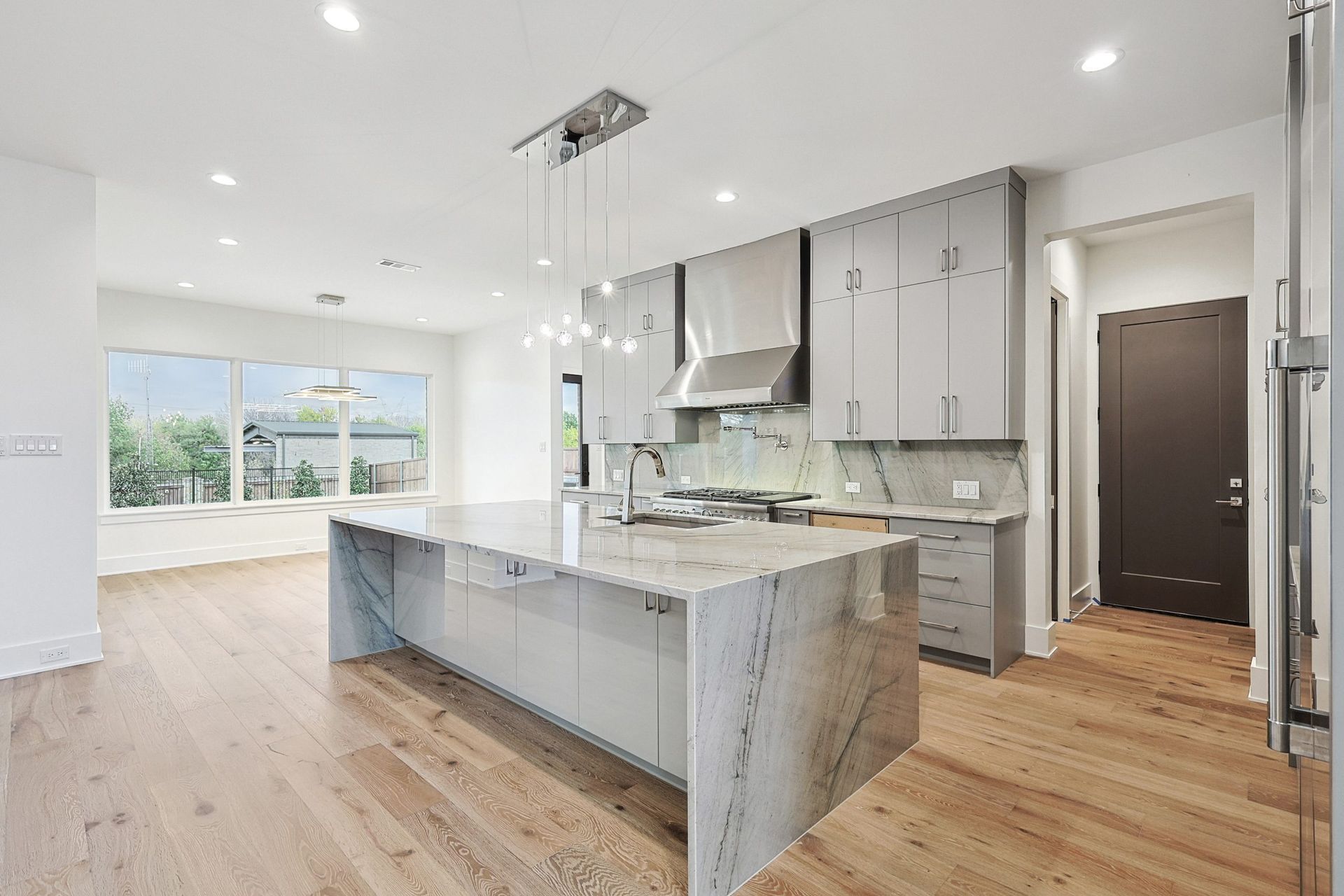 A modern kitchen with a large marble island, gray cabinetry, stainless steel appliances, and light wood flooring.