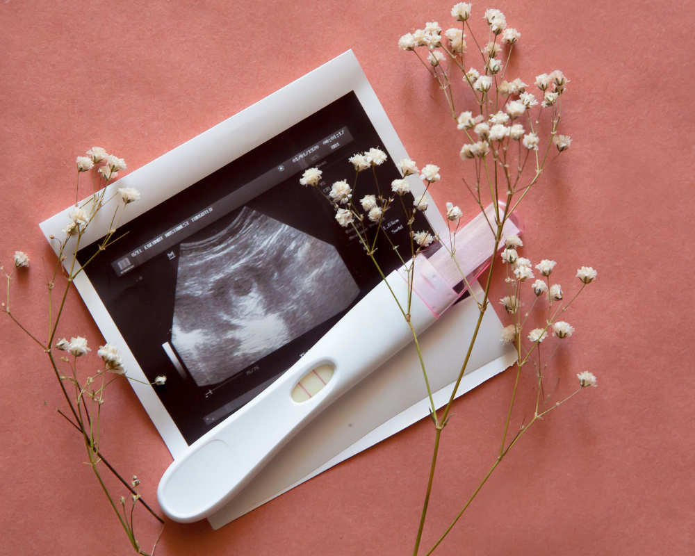 Pregnancy test and ultrasound photo with baby's image, pink background and white flowers.
