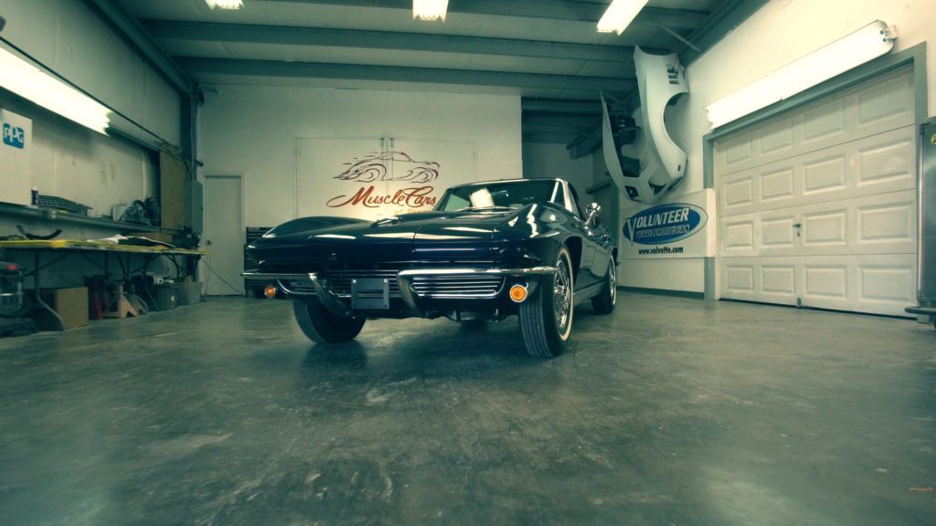 Black vintage car in a garage with tools, a logo on the wall, and a partial car body hanging.