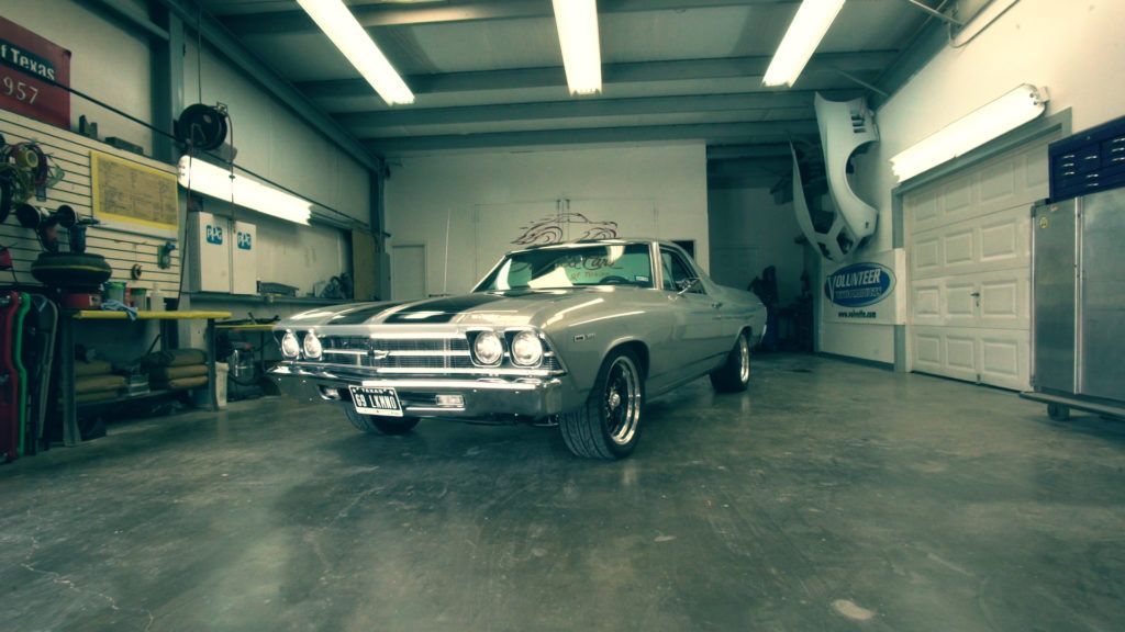 Silver classic El Camino in a well-lit garage, black racing stripes, tools and equipment visible.