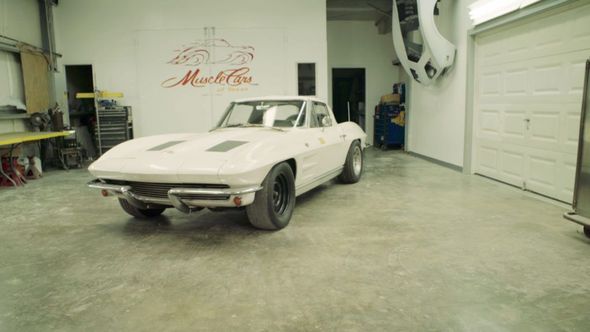 White vintage Corvette in a garage, with a suspended car in the background.