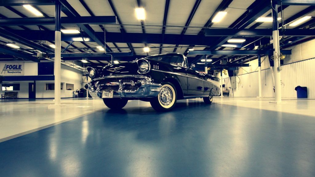 Black classic car parked in a large warehouse, white-wall tires, blue floor.