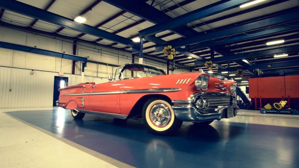 Red classic convertible car parked inside a well-lit garage with blue and white accents.