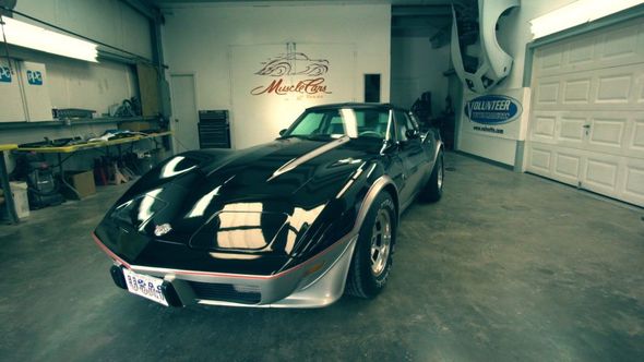 Black Corvette in a garage, silver trim, under bright lights.