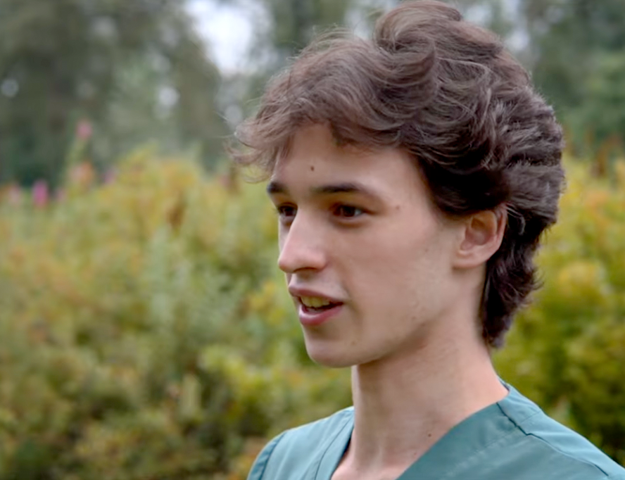 Young person with curly brown hair in a green top, outdoors, talking.