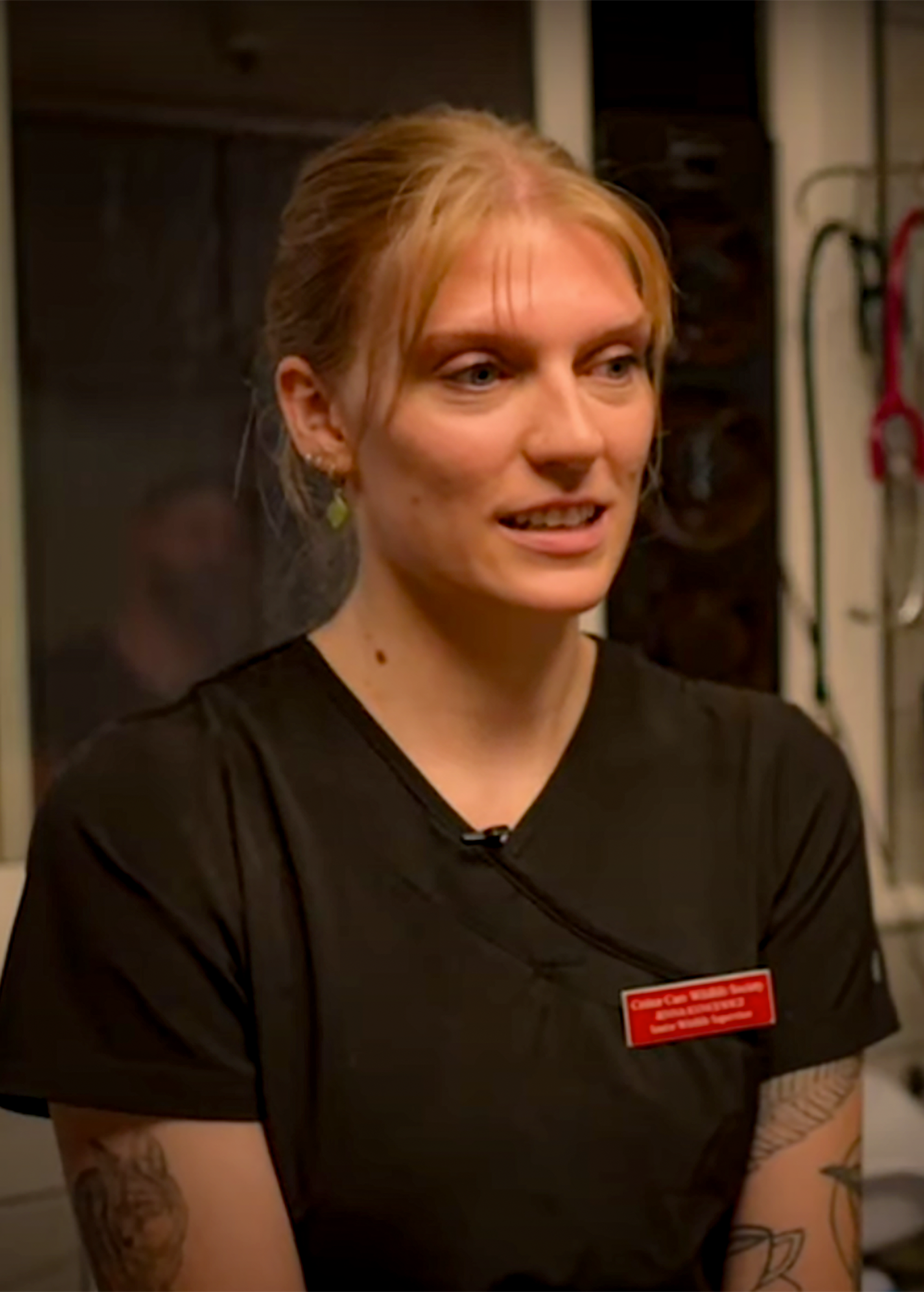 Woman in black scrubs with a name tag, in a medical setting. She has blonde hair and a tattoo.