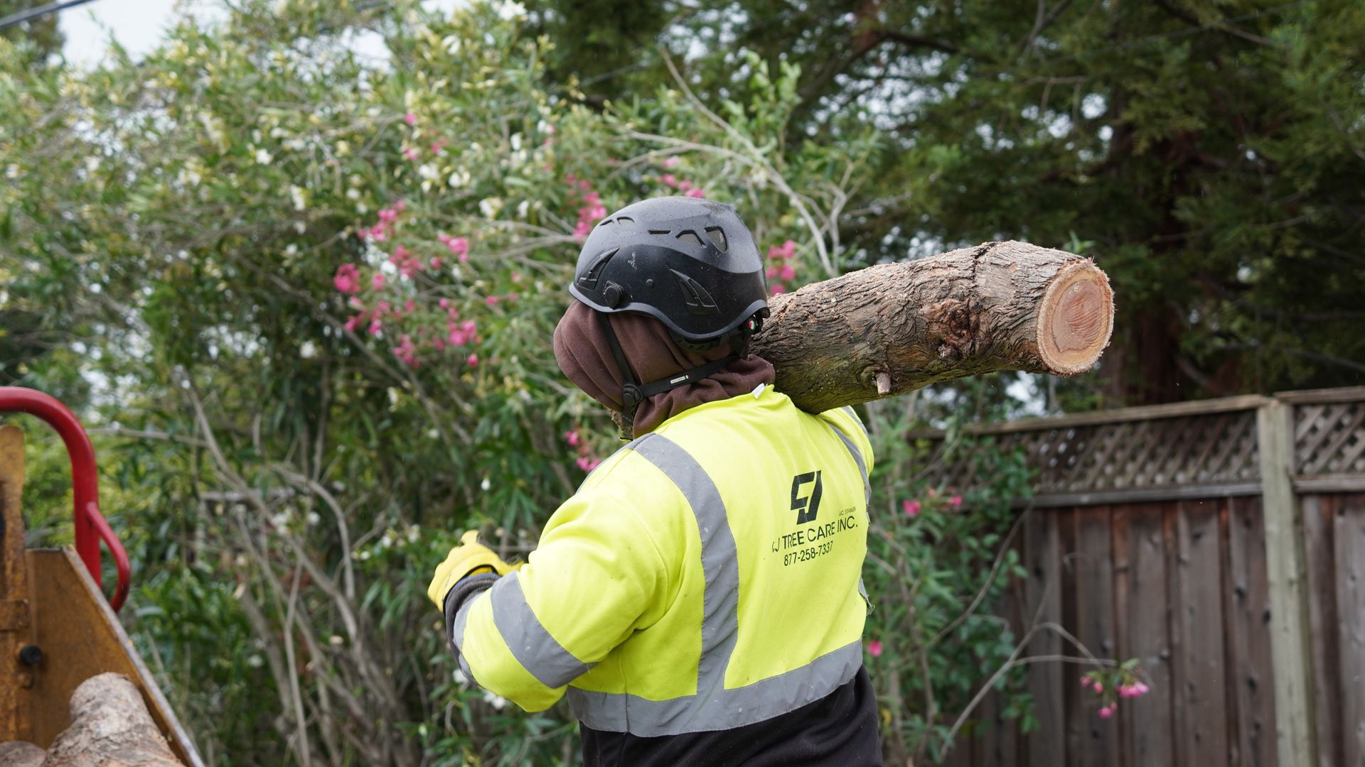 Worker in high-vis jacket and helmet carrying a large log in a garden or yard.