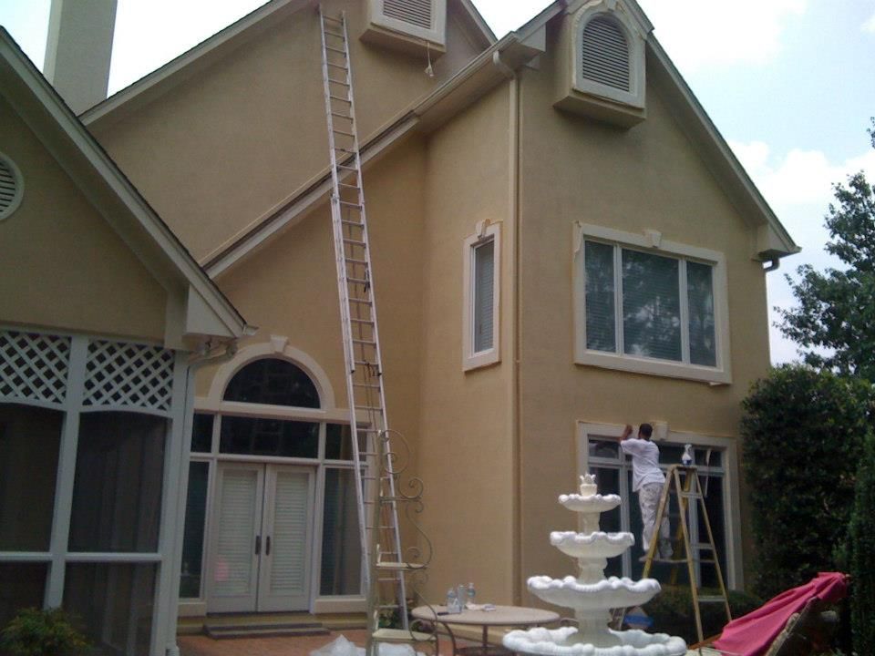House being painted; a painter on a ladder near a window. Tall ladder against the side of the building. Beige house.