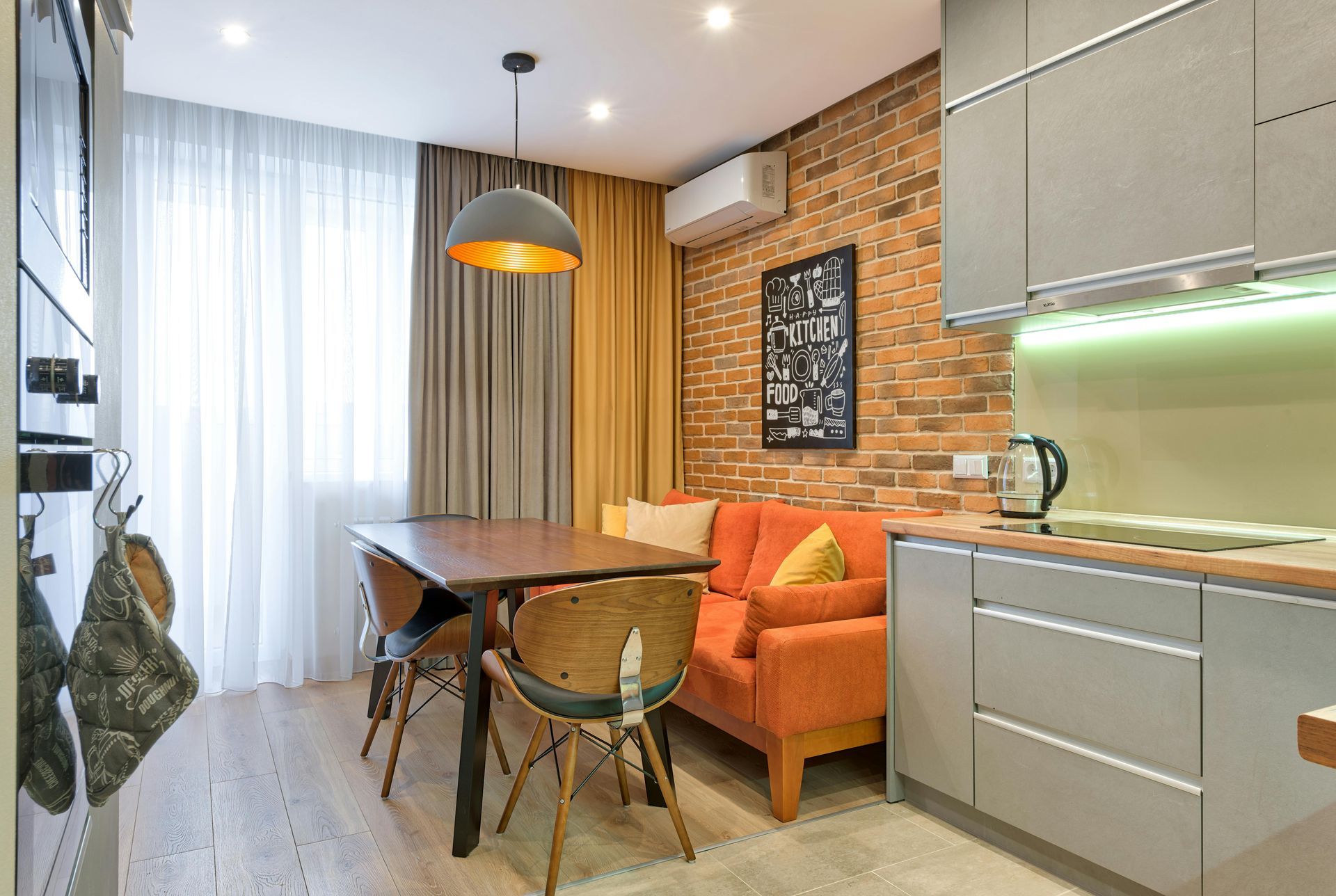 Small kitchen with orange sofa, dining table, exposed brick wall, and grey cabinetry.