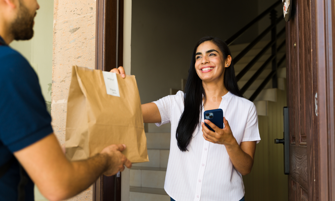 Smiling delivery worker hands a package to a person at the doorway, holding a phone.