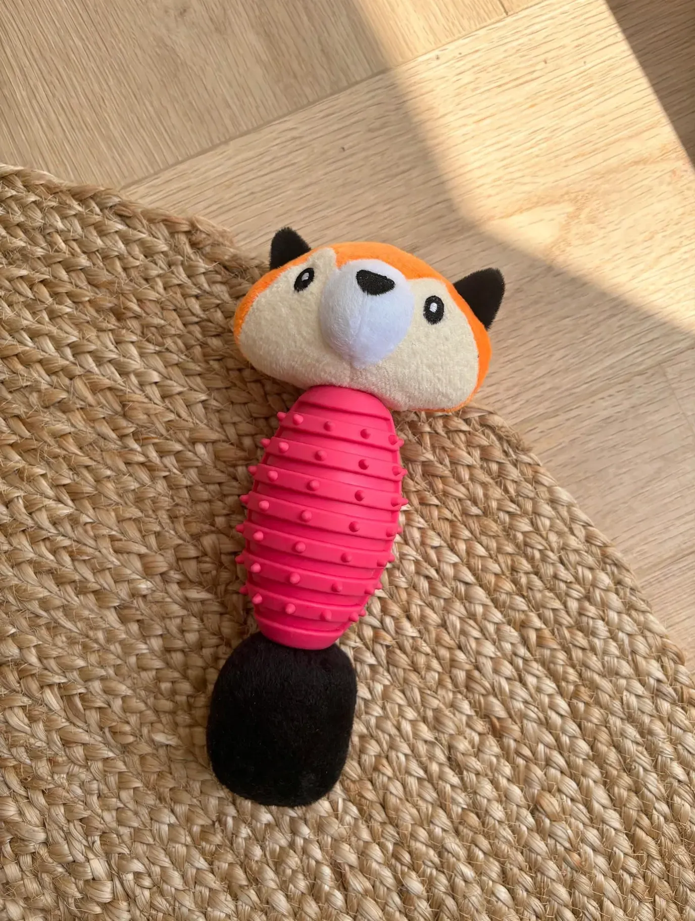 Pequeño juguete de peluche para gatos con cuerpo rosa y cola negra sobre una alfombra tejida bajo la luz del sol.