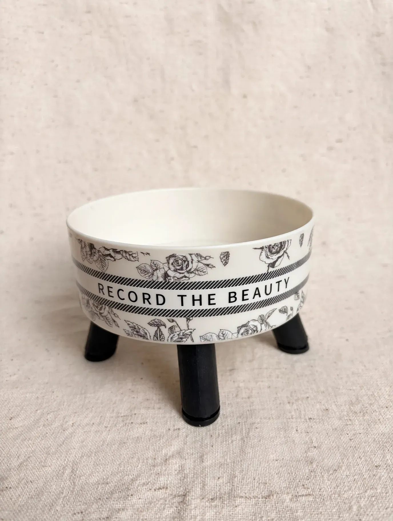 Bol decorativo blanco sobre patas negras con banda estampada y texto parcial sobre fondo de tela beige.