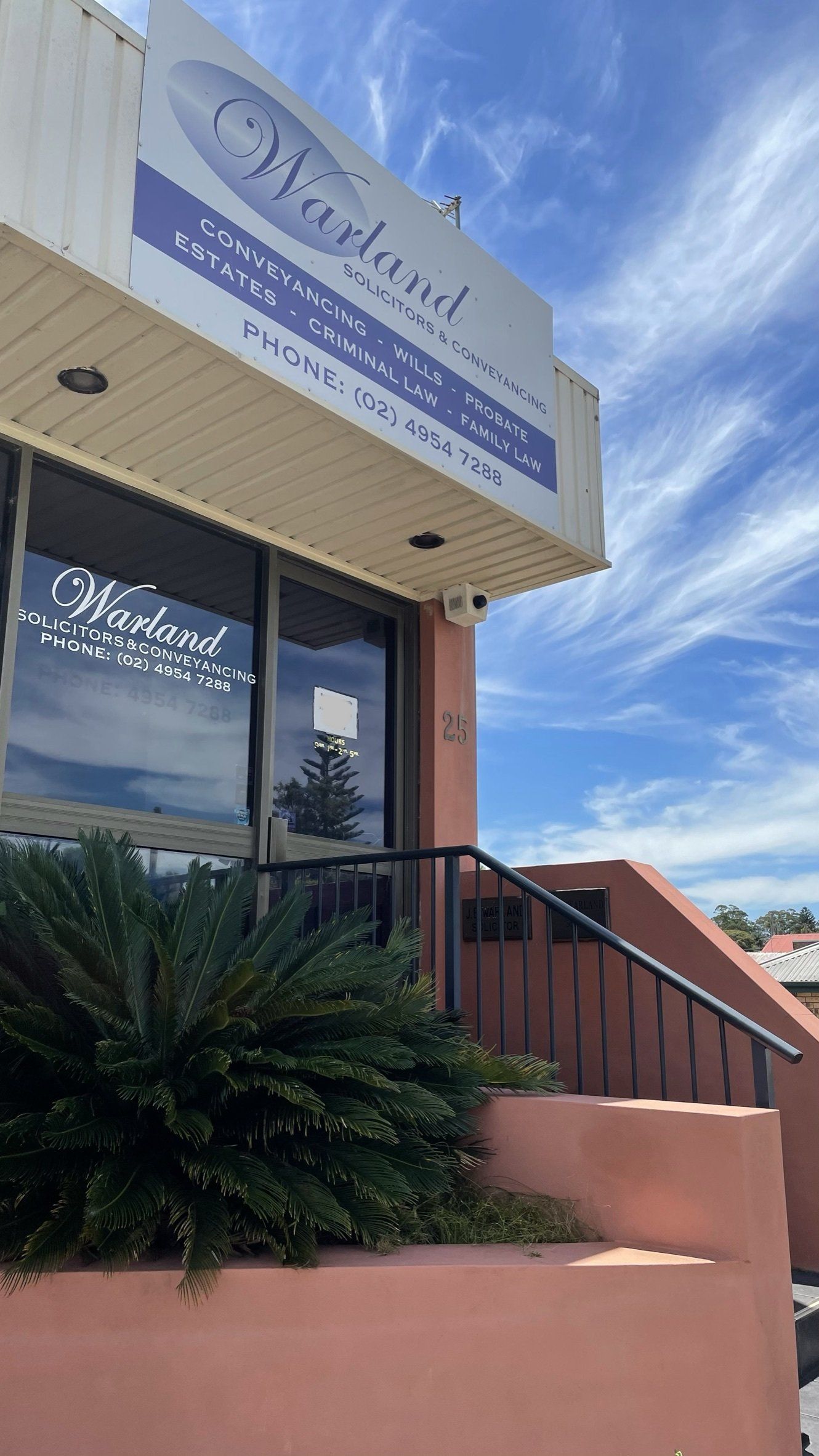 Storefront with a blue-and-white sign, glass door, pink steps, and a palm plant under a clear sky - Warland Solicitors & Conveyancing