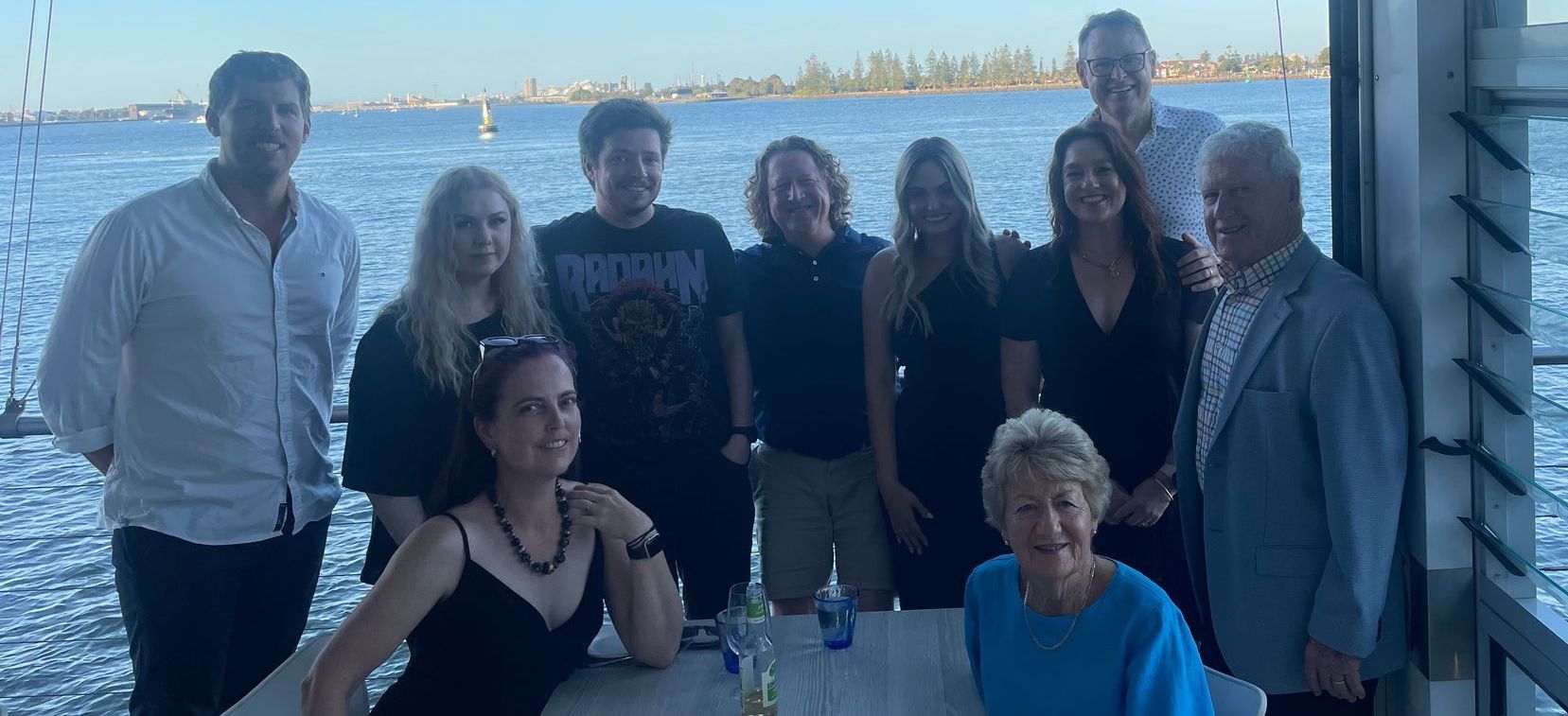 Group of people posing on a waterfront deck with water and a distant shoreline behind them.