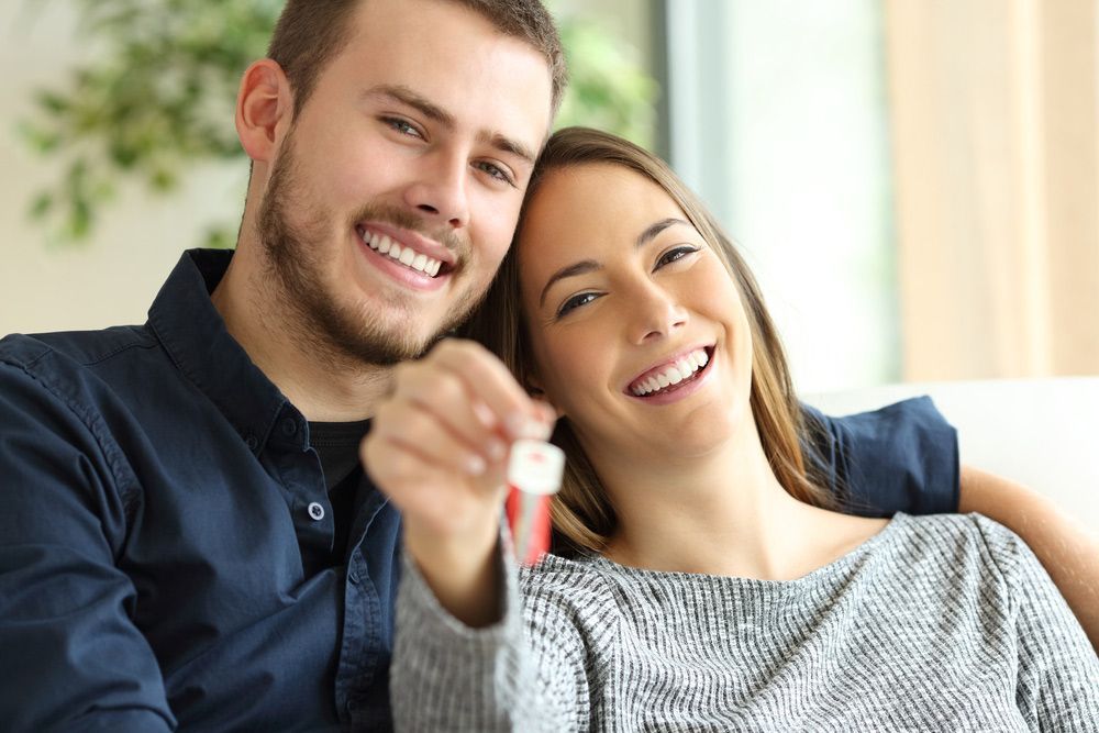 Smiling couple on a couch holding out a key in a bright living room