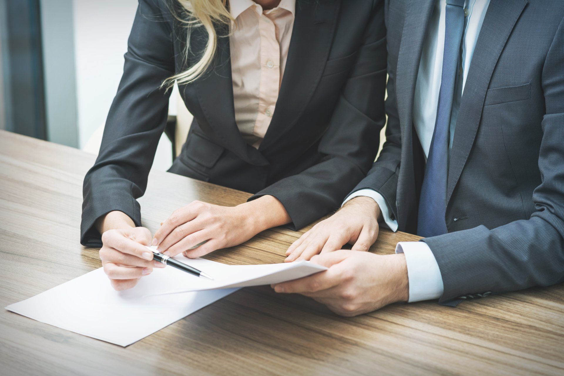 Two professionals in suits reviewing documents at a desk, one signing paperwork with a pen.