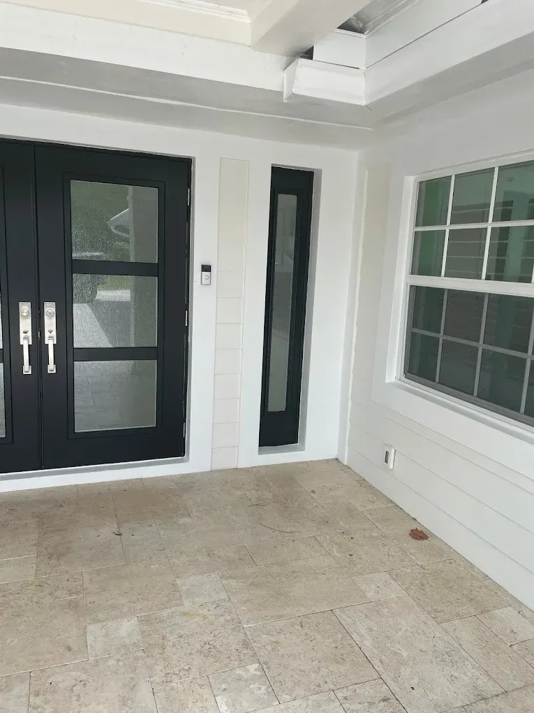 Covered porch with black double doors, narrow side window, and white siding next to a large window.