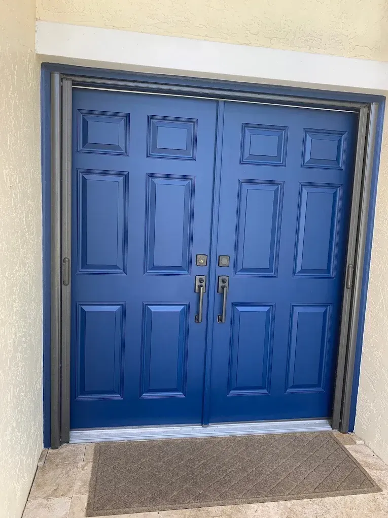 Blue double front doors with brass handles and a gray doormat in a beige exterior wall.