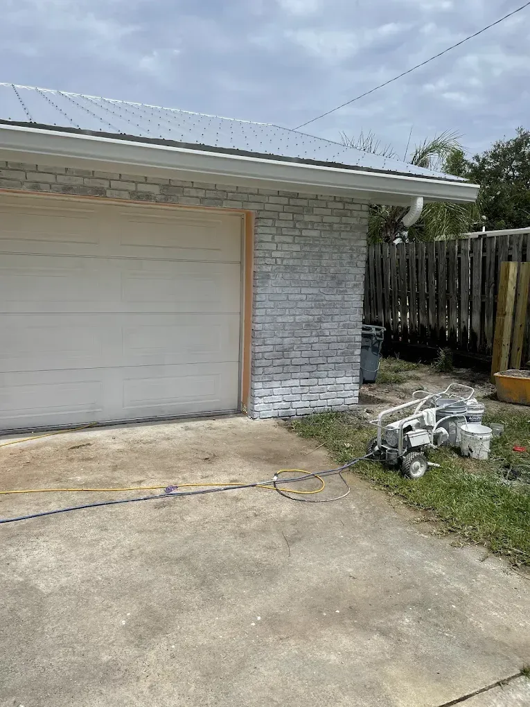White garage with closed door, brick wall, driveway, and a small pile of debris beside a wooden fence.