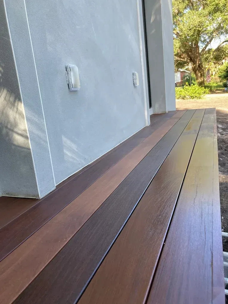 Wooden deck boards beside a gray house wall, with sunlight and trees in the background